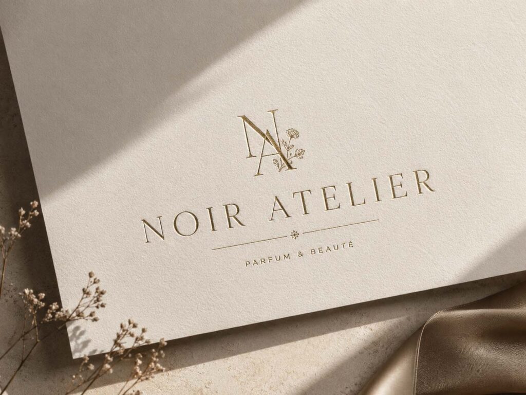 Elegantes Noir-Atelier-Logo in Gold auf strukturiertem cremefarbenem Papier mit floralen Details.